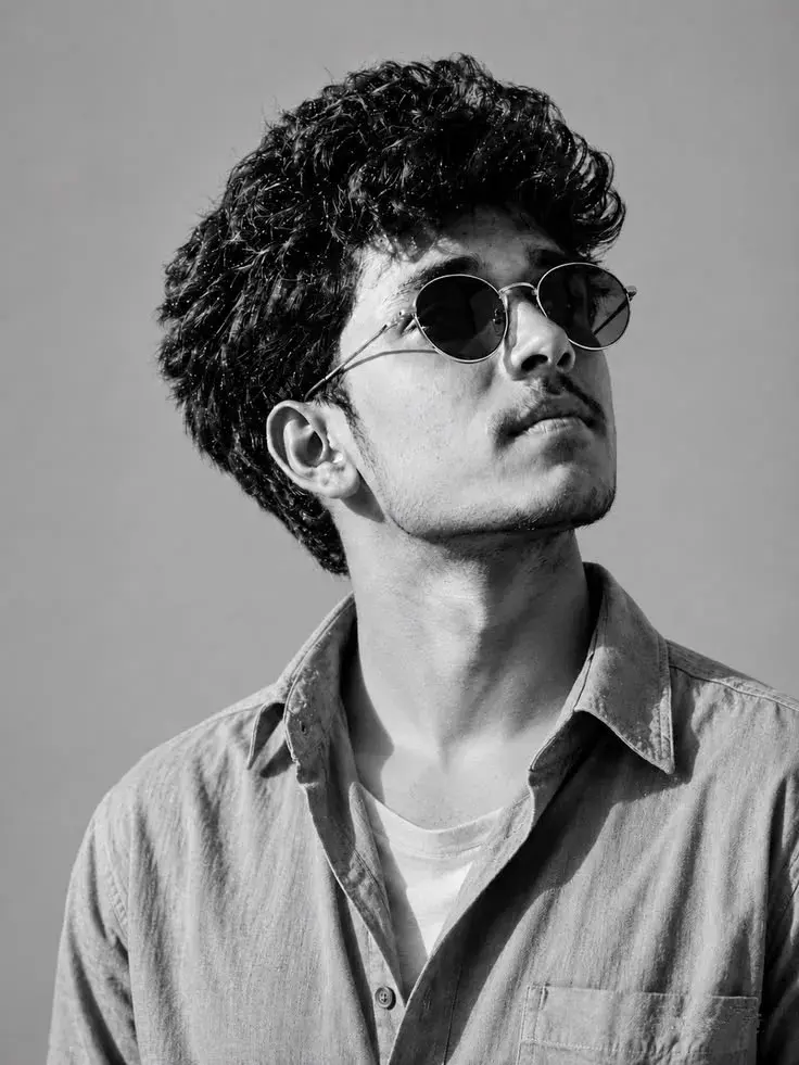 Black-And-White Low-Angle Close-Up Portrait Of Young South Asian Man With Curly Crop And Round Sunglasses Looking Upward