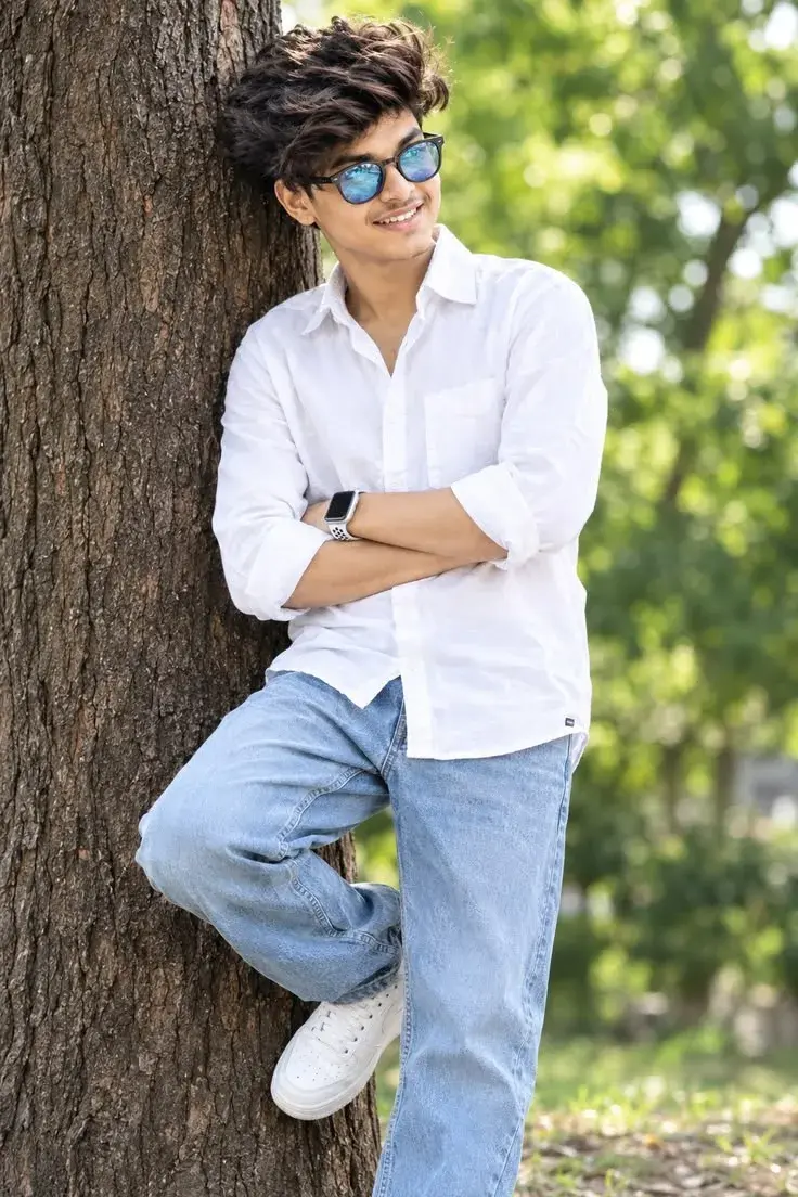 Candid Full-Length Outdoor Portrait Of Smiling Young South Asian Man Leaning On Tree Trunk In White Linen Shirt