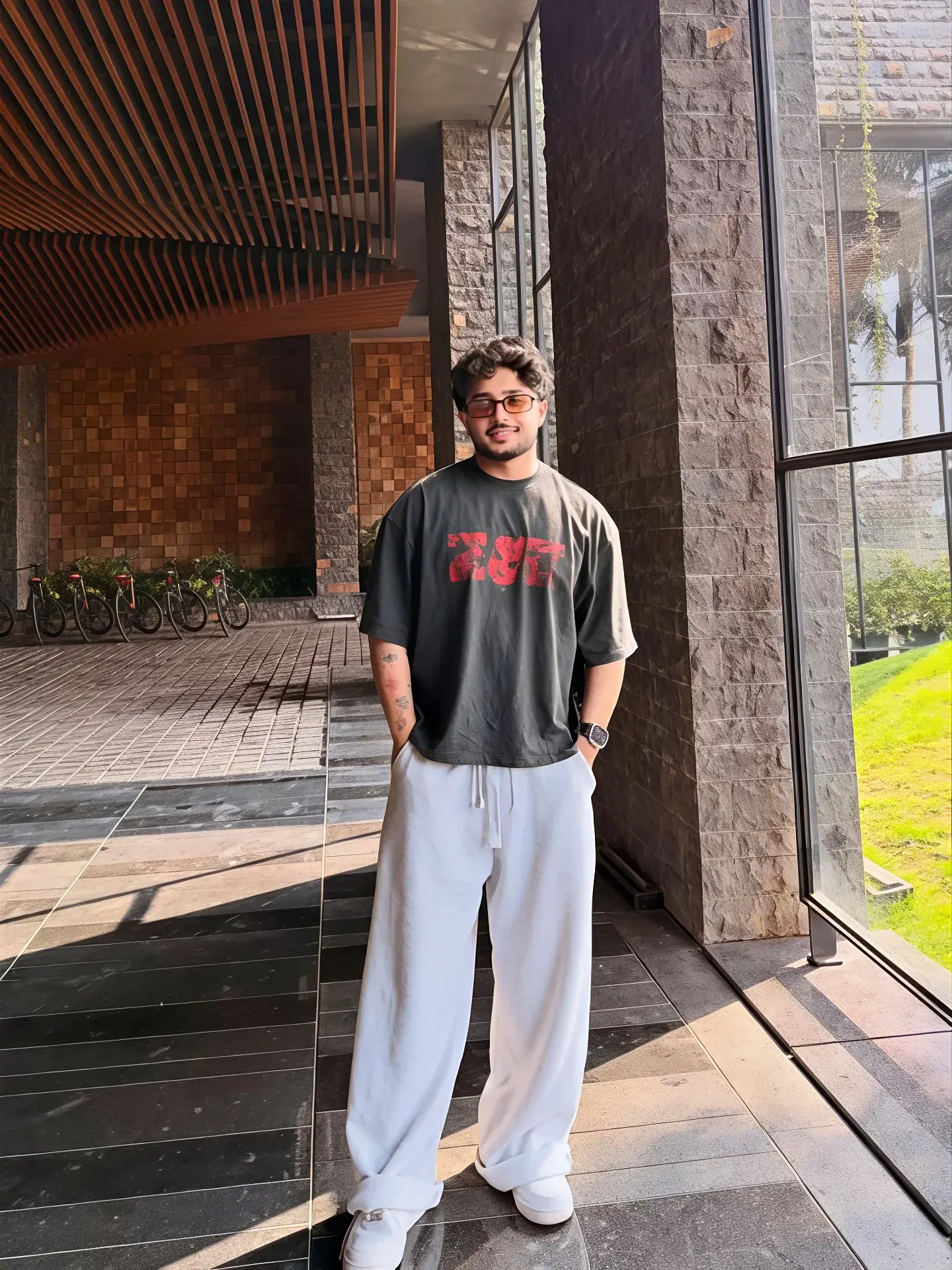 Candid Full-Length Portico Portrait Of Young South Asian Man In Dark Graphic T-Shirt And White Baggy Sweatpants
