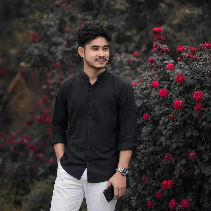 Candid Garden Portrait Of Smiling Young South Asian Man In Black Mandarin Shirt And White Trousers Beside Red Rose Bushe