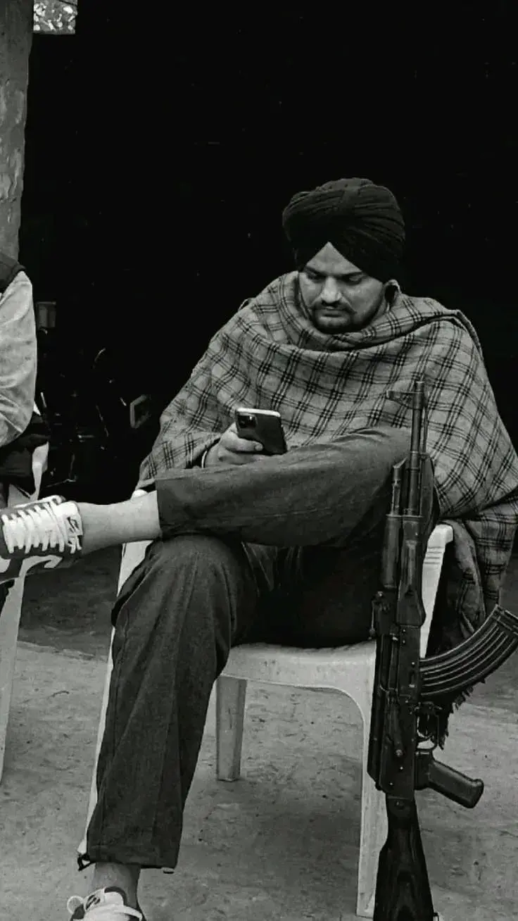 Candid Gritty Monochrome Portrait Of South Asian Man In Turban And Plaid Shawl Seated With Rifle And Smartphone