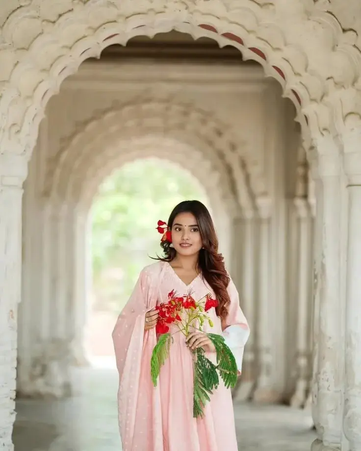 Candid Heritage Archway Portrait Of Serene Young South Asian Woman In Pastel Pink Anarkali Holding Red Flowers