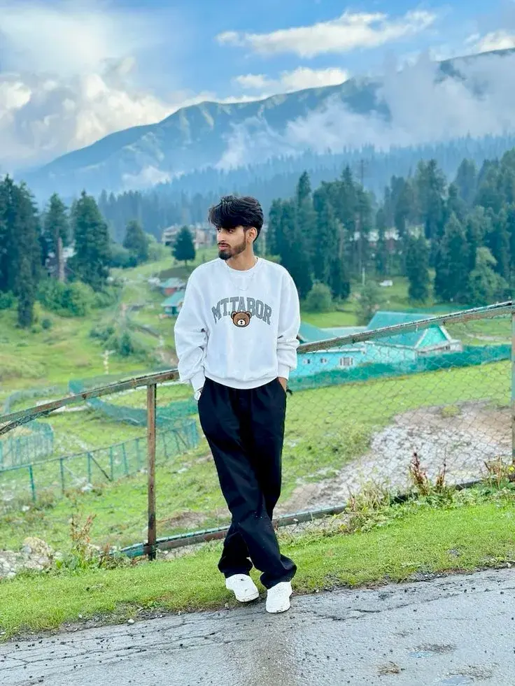Candid Himalayan Valley Roadside Portrait Of Young South Asian Man In White Teddy Graphic Sweatshirt Leaning On Wire Fen