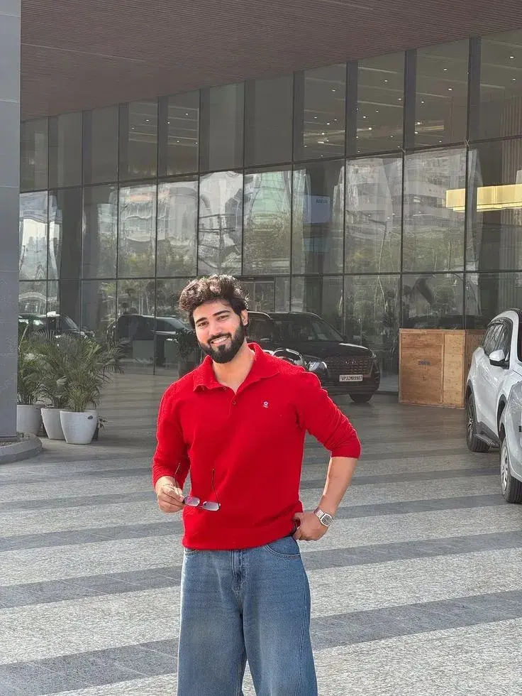 Candid Hotel Entryway Portrait Of Smiling Young South Asian Man In Red Knit Polo And Baggy Denim Jeans Holding Sunglasse