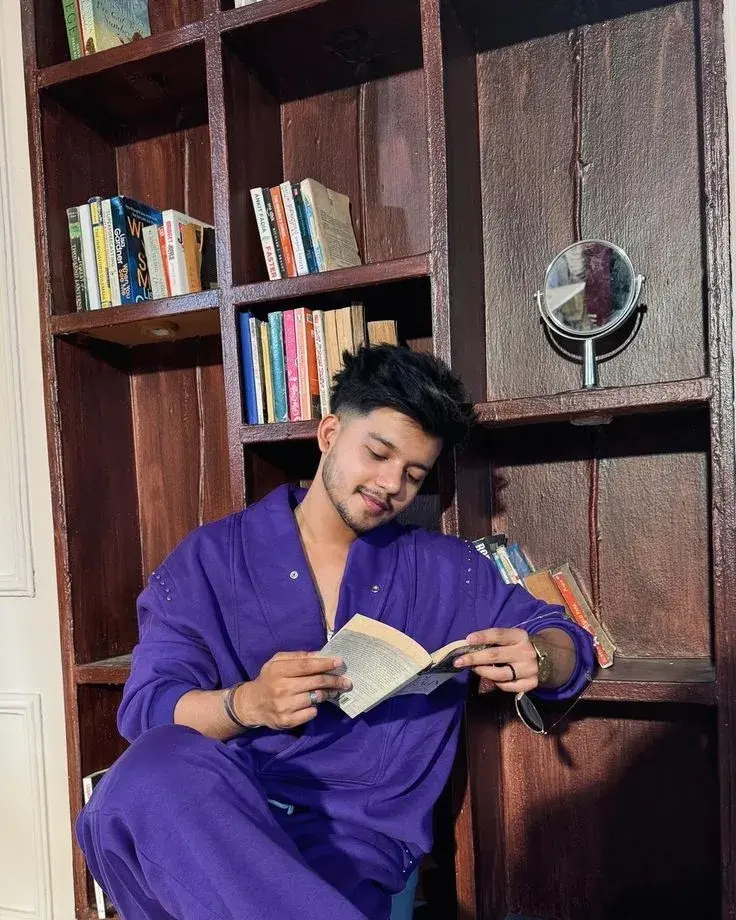Candid Indoor Bookshelf Nook Portrait Of Smiling Young South Asian Man In Purple Tracksuit Reading Paperback Book