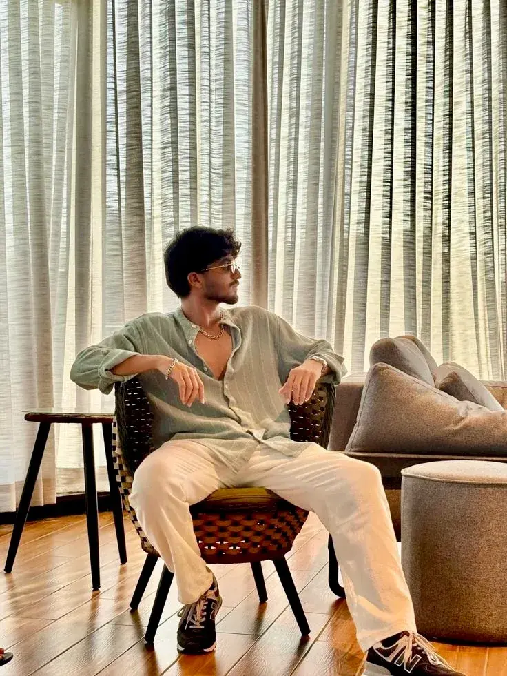 Candid Indoor Lounge Portrait Of Young South Asian Man In Mint Striped Shirt And White Trousers Seated On Woven Armchair