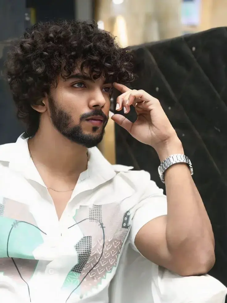 Candid Indoor Portrait Of Thoughtful Young South Asian Man With Curly Hair And Silver Watch Resting Hand Near Temple