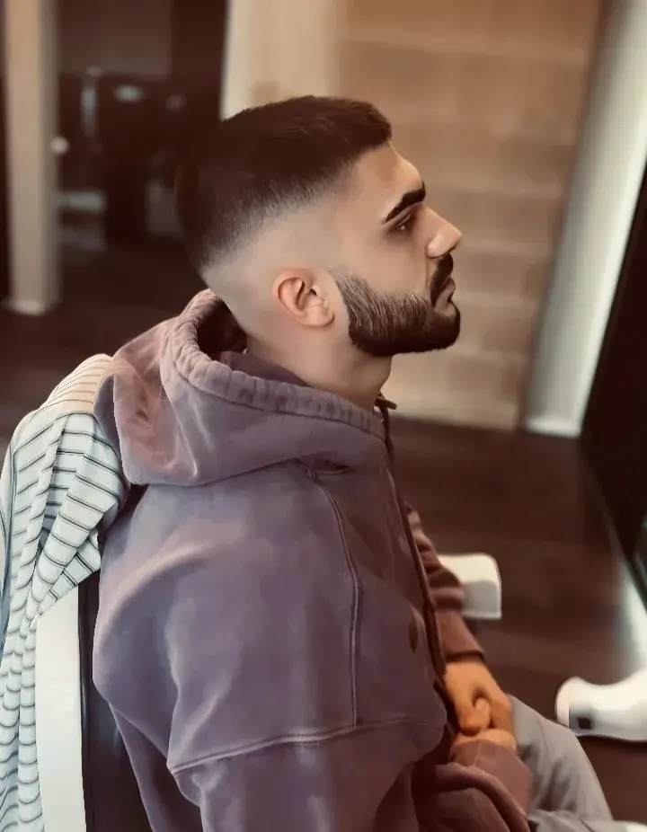 Candid Indoor Side Profile Portrait Of Young South Asian Man With Crisp High Skin Fade And Contoured Boxed Beard In Mauv