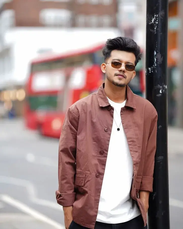 Candid London Street Portrait Of Young South Asian Man In Terracotta Overshirt And Tinted Sunglasses Leaning Against Lam