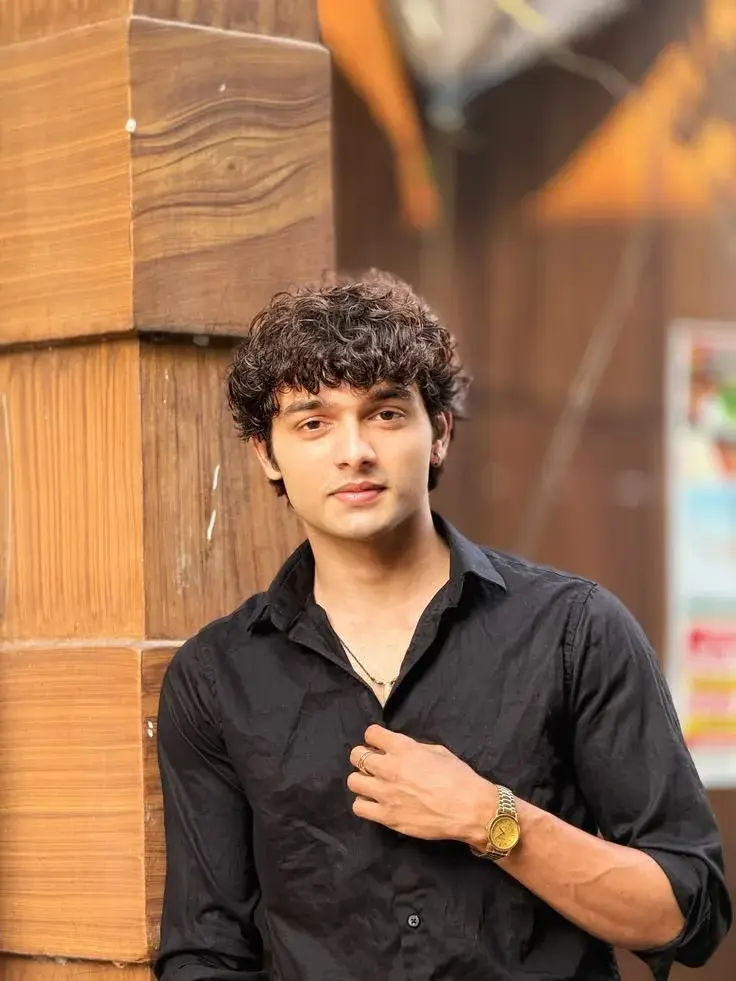 Candid Medium-Shot Outdoor Portrait Of Young South Asian Man With Curly Bangs In Black Shirt Leaning Against Wood Pillar