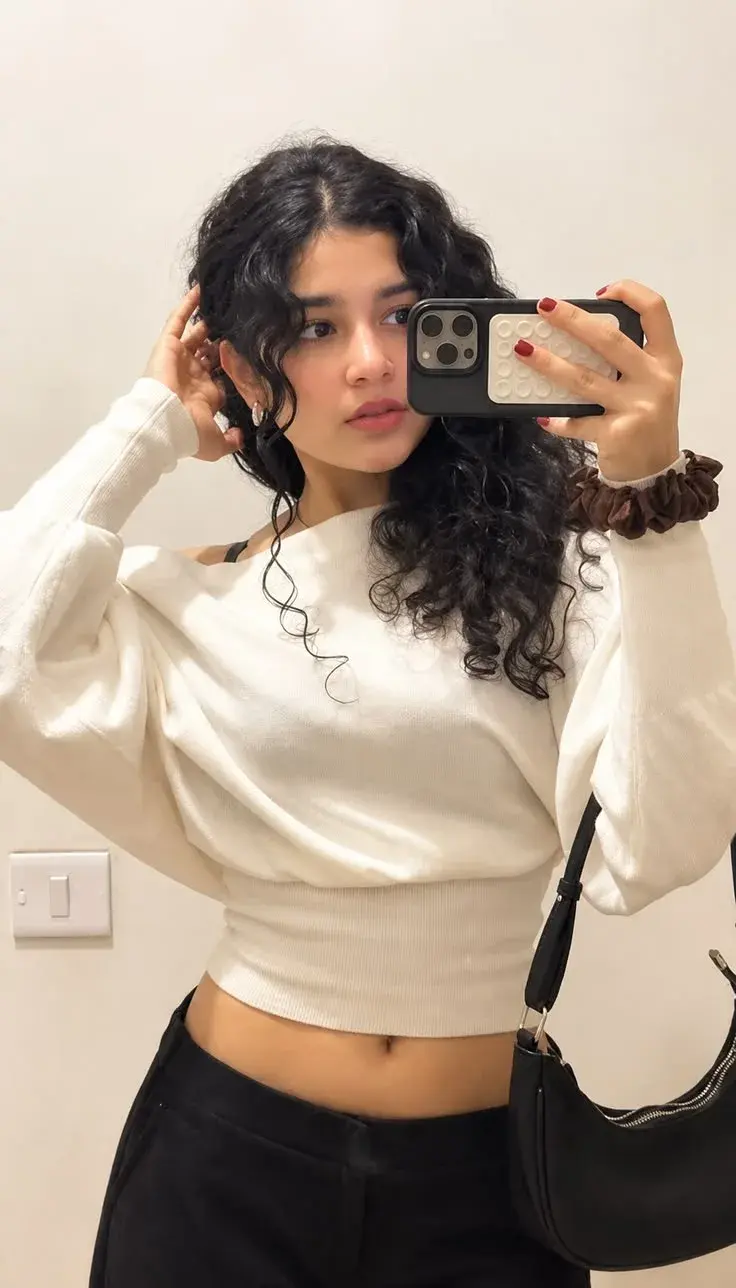 Candid Mirror Selfie Portrait Of Young South Asian Woman With Curly Hair In Off-Shoulder Cream Sweater And Black Trouser