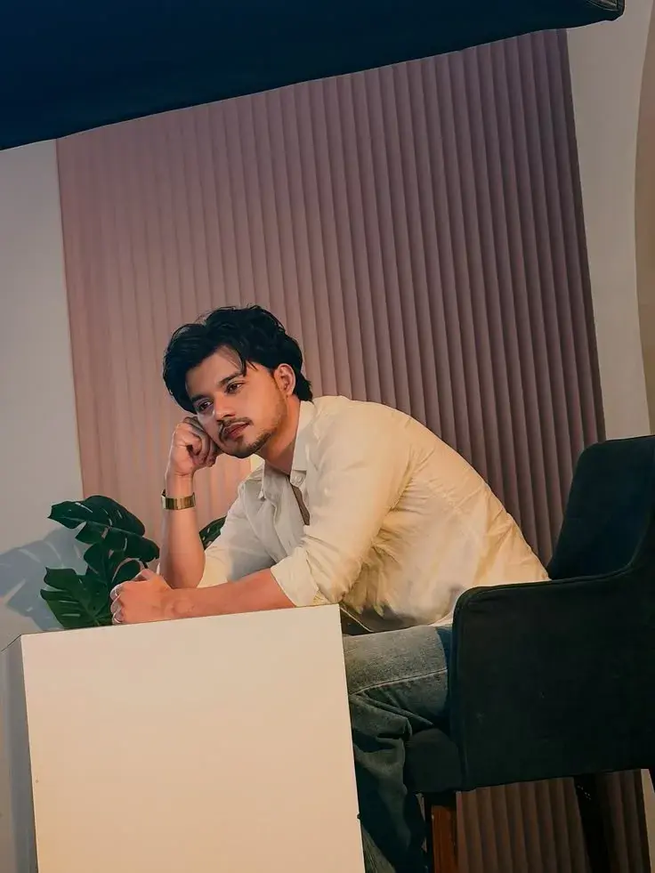 Candid Modern Studio Portrait Of Young South Asian Man In Off-White Linen Shirt Leaning On White Cube By Fluted Wall
