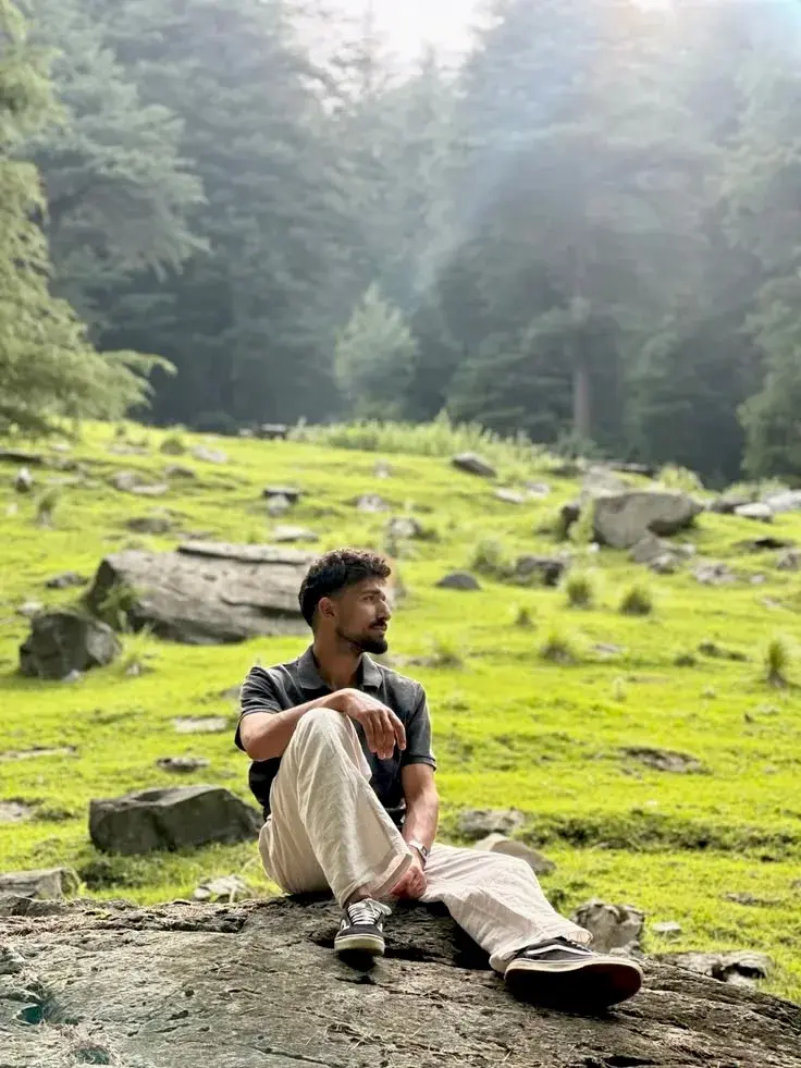 Candid Mountain Meadow Portrait Of Young South Asian Man Sitting On Weathered Rock In Sunlit Pine Valley