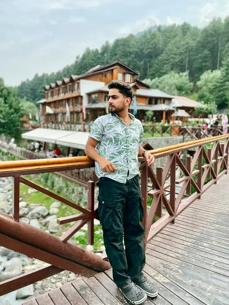 Candid Mountain Valley Bridge Portrait Of Young South Asian Man In Green Botanical Shirt Leaning On Wooden Railing