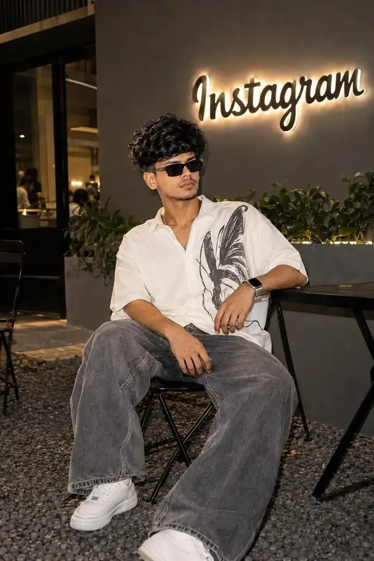 Candid Night Cafe Portrait Of Young South Asian Man With Curly Hair In Graphic Shirt And Baggy Jeans Under Glowing Insta