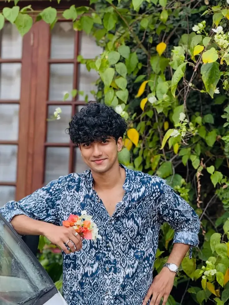 Candid Outdoor Garden Portrait Of Young South Asian Man In Blue Ikat Shirt Leaning On Car With Flowers