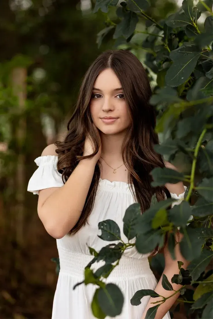 Candid Outdoor Garden Portrait Of Young Woman In White Off-Shoulder Smocked Dress Framed By Greenery