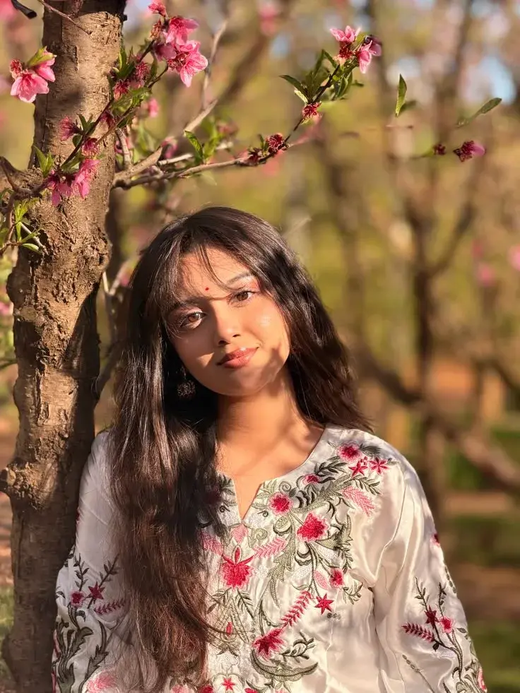 Candid Outdoor Golden Hour Portrait Of Young South Asian Woman In Embroidered Kurti Leaning On Blooming Peach Blossom Tr