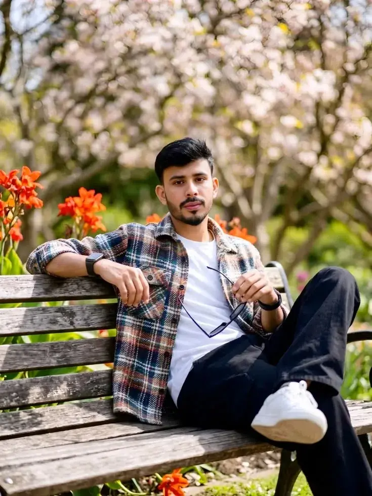 Candid Outdoor Park Portrait Of Young South Asian Man In Plaid Overshirt Relaxing On Wooden Bench With Sunglasses