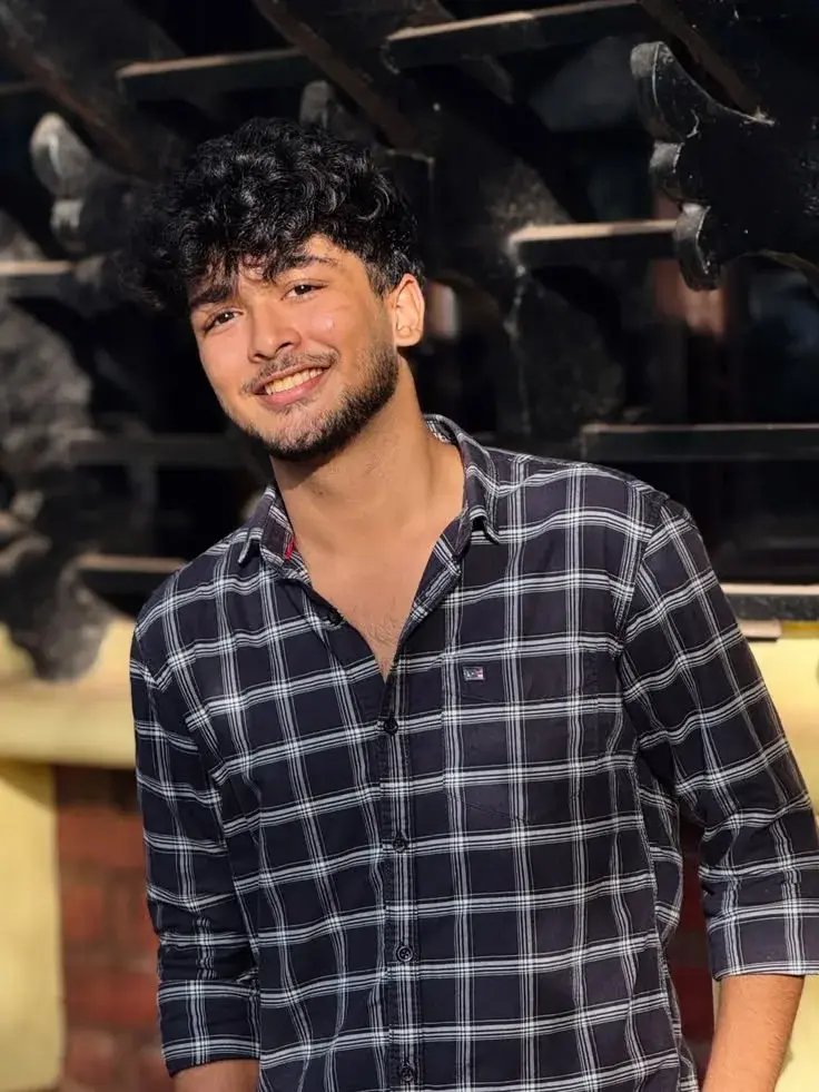 Candid Outdoor Portrait Of Smiling Young South Asian Man In Black Plaid Shirt Against Dark Trellis