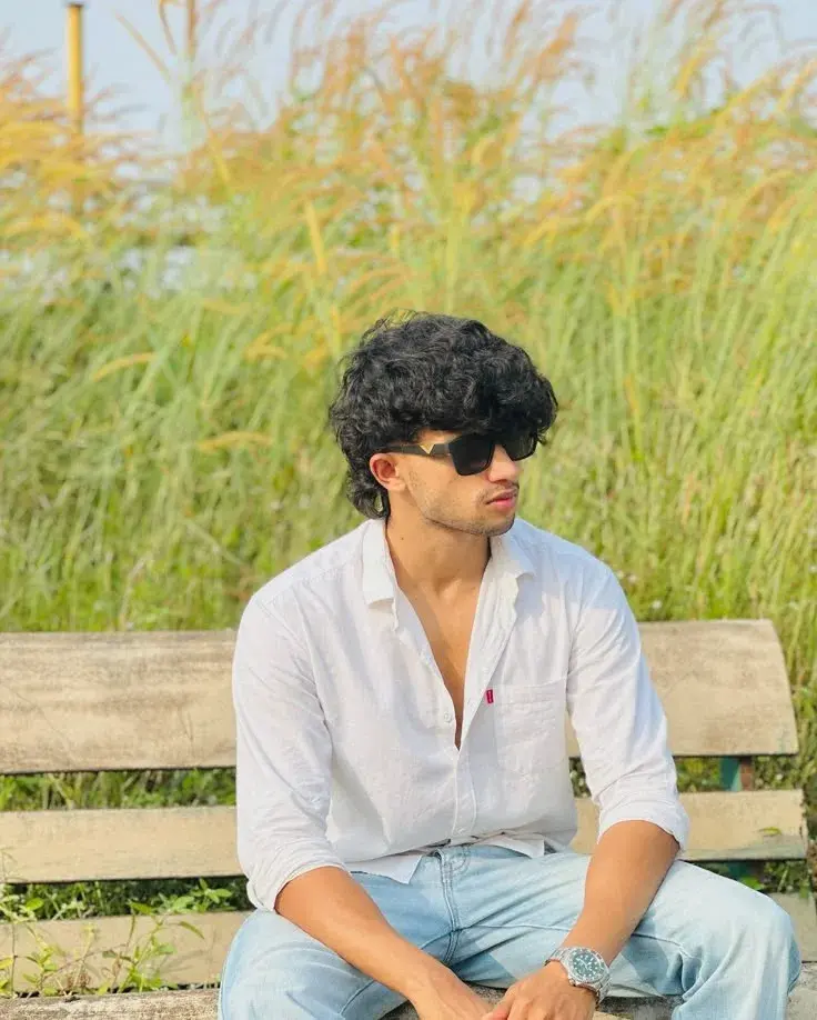 Candid Outdoor Portrait Of Young Indian Man Seated On Bench With Wild Grass