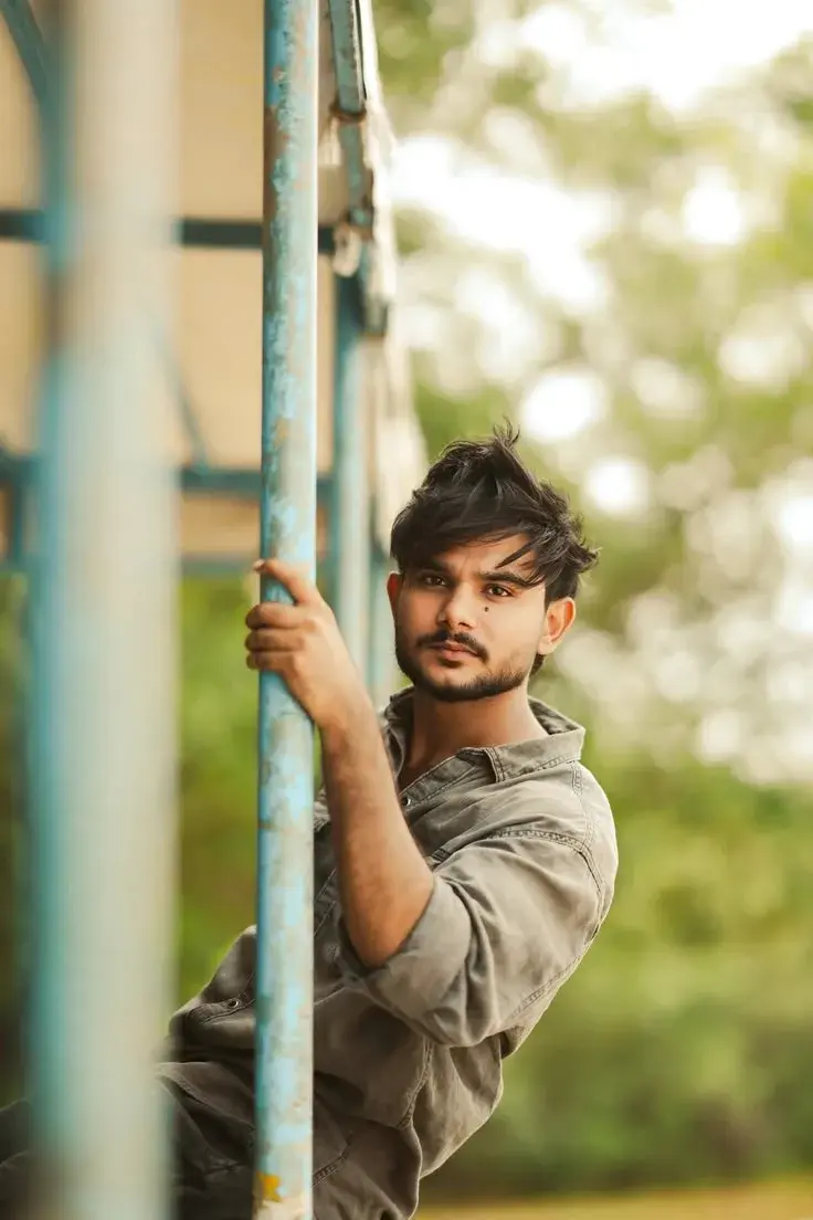 Candid Outdoor Portrait Of Young South Asian Man Holding Blue Metal Pole With Bokeh Foliage