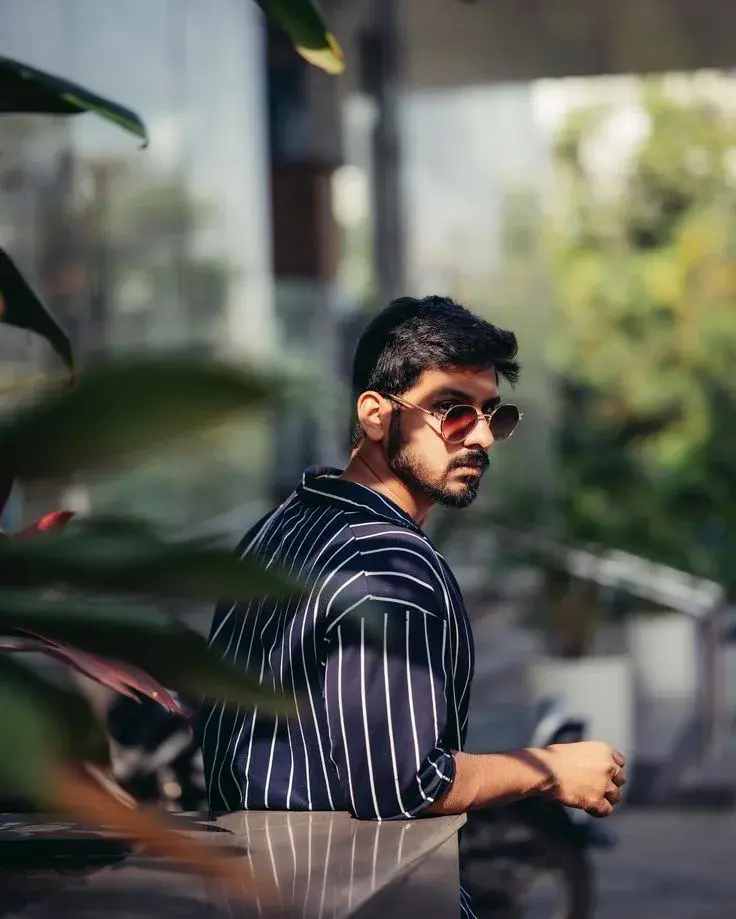 Candid Outdoor Portrait Of Young South Asian Man In Navy Striped Shirt And Retro Sunglasses Leaning On Ledge