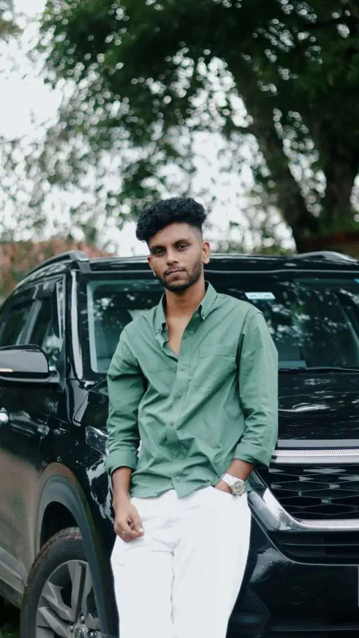 Candid Outdoor Portrait Of Young South Asian Man Leaning On Black Suv In Sage Green Shirt And White Trousers