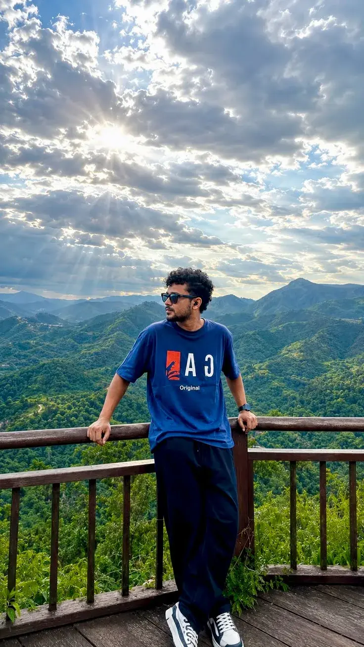 Candid Outdoor Viewpoint Portrait Of Young South Asian Man Leaning On Wooden Balcony Railing With Mountain Ridge And Dra