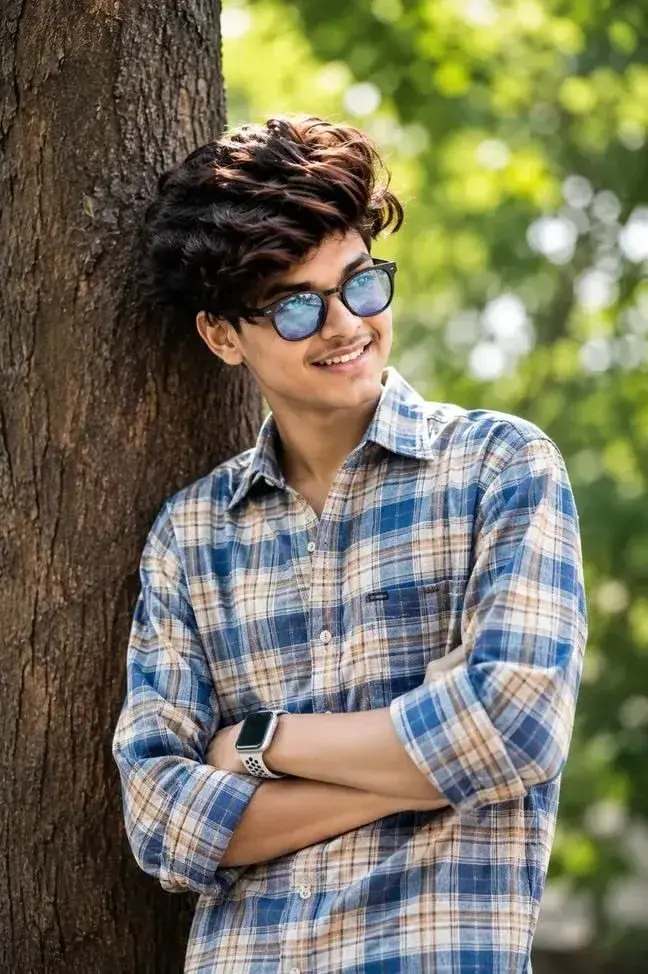 Candid Outdoor Waist-Up Portrait Of Smiling Young Indian Man Leaning On Tree Trunk