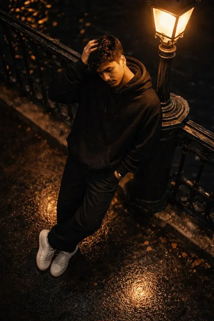Candid Rainy Night Canal Bridge Portrait Of Young South Asian Man In Black Hoodie Leaning Against Vintage Streetlamp