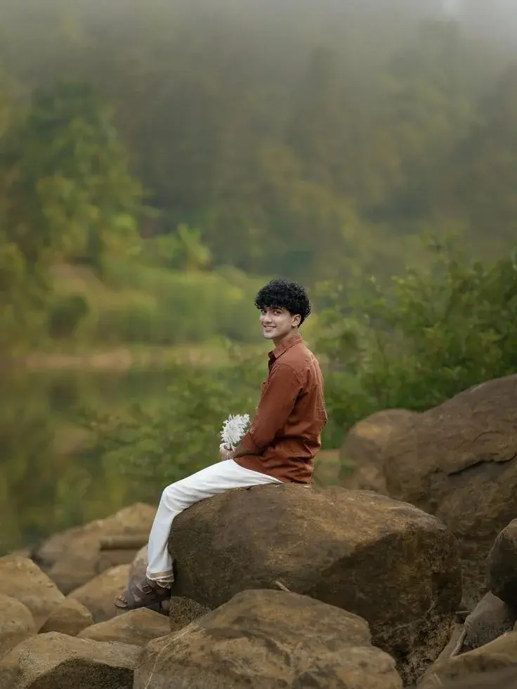 Candid Riverside Nature Portrait Of Smiling Young South Asian Man With Curly Fringe In Rust Shirt And Dhoti Seated On Bo
