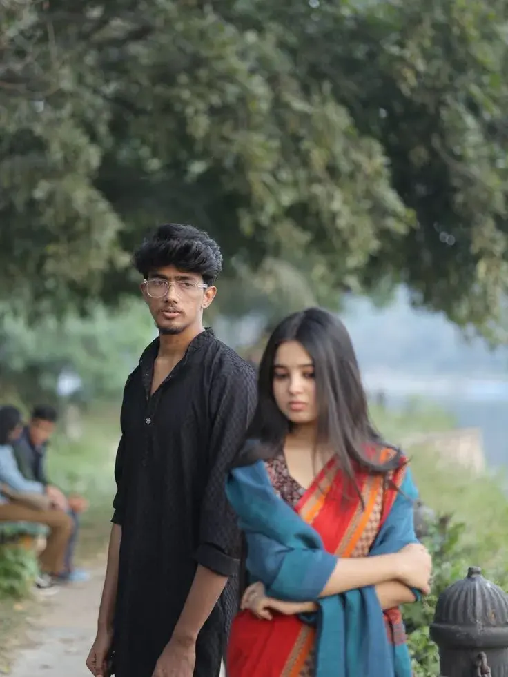 Candid Riverside Promenade Portrait Of Pensive Young South Asian Couple With Woman In Red Saree And Man In Black Kurta