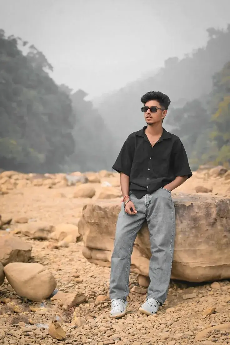 Candid Rocky Valley Riverbed Portrait Of Young South Asian Man In Oversized Black Shirt And Sunglasses Leaning On Boulde