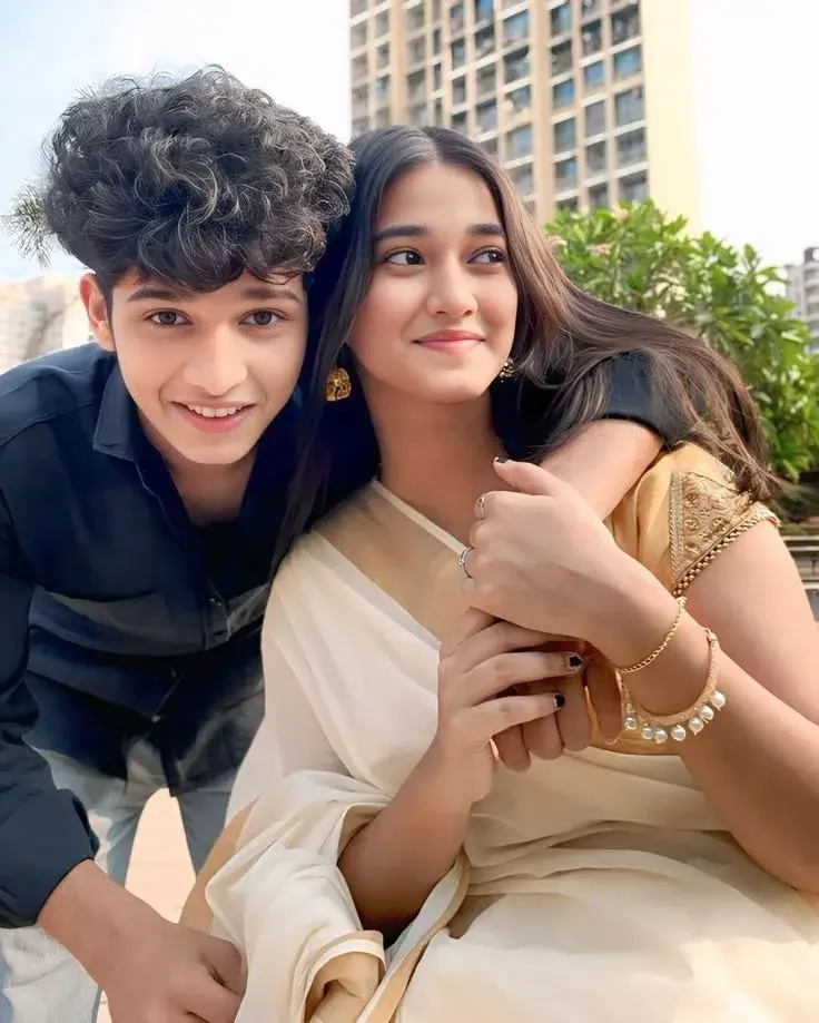 Candid Rooftop Portrait Of Smiling Young South Asian Boy Leaning Over Older Sister In Traditional White Kasavu Saree