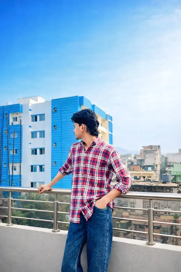 Candid Rooftop Terrace Portrait Of Young South Asian Man With Wavy Shag Hair In Red Plaid Flannel Shirt Leaning On Raili