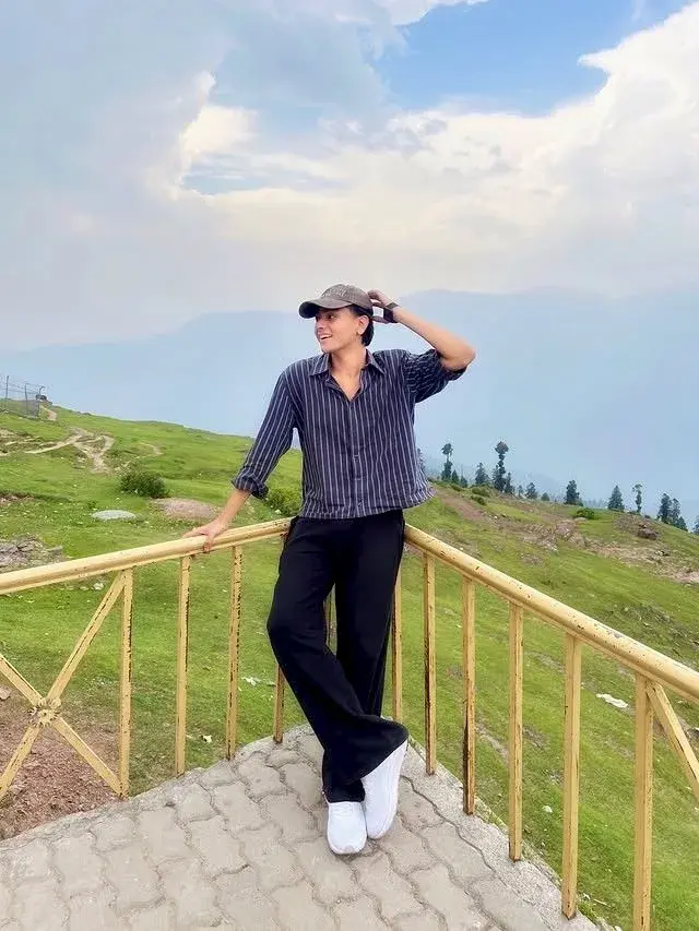 Candid Scenic Mountain Meadow Overlook Portrait Of Smiling Young South Asian Man In Striped Shirt And Cap Leaning On Yel