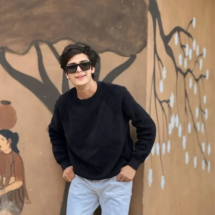 Candid Square Portrait Of Smiling Young South Asian Man In Black Knit Sweater And Flat-Top Sunglasses Leaning On Painted
