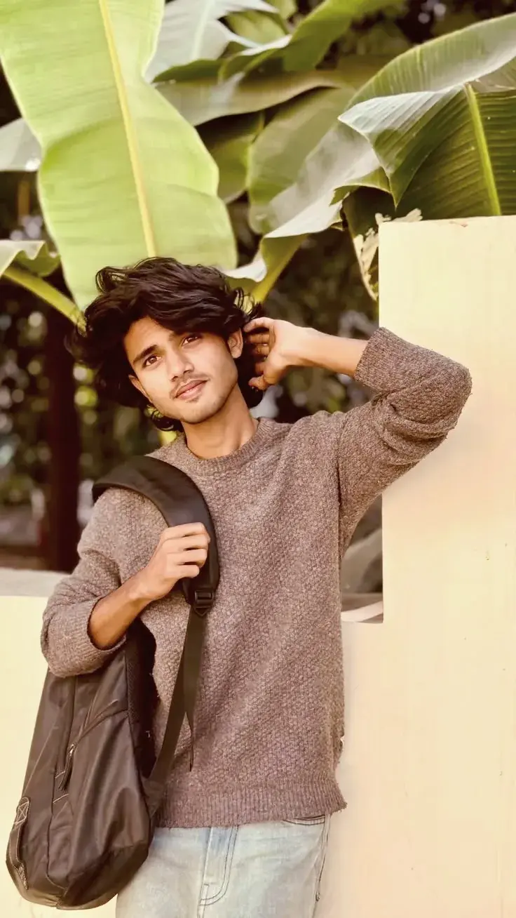 Candid Sunlit Banana Grove Portrait Of Young South Asian Man With Flowing Wavy Hair In Brown Knit Sweater Holding Backpa