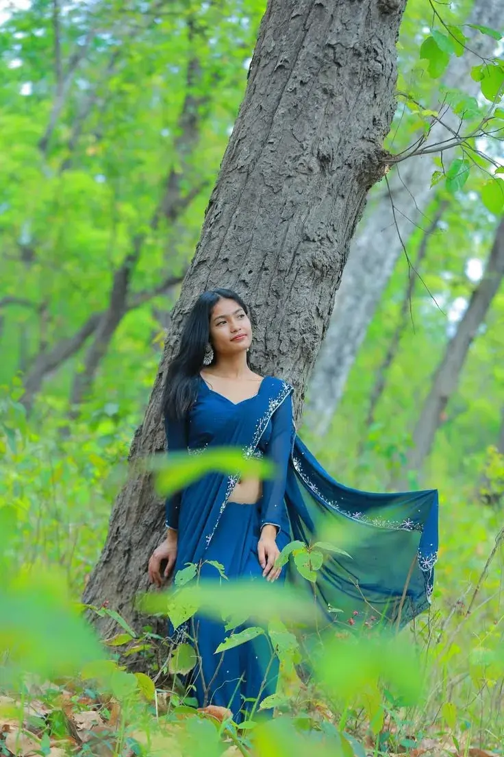 Candid Sunlit Forest Portrait Of Young South Asian Woman In Blue Georgette Saree Leaning Against Mossy Tree Trunk
