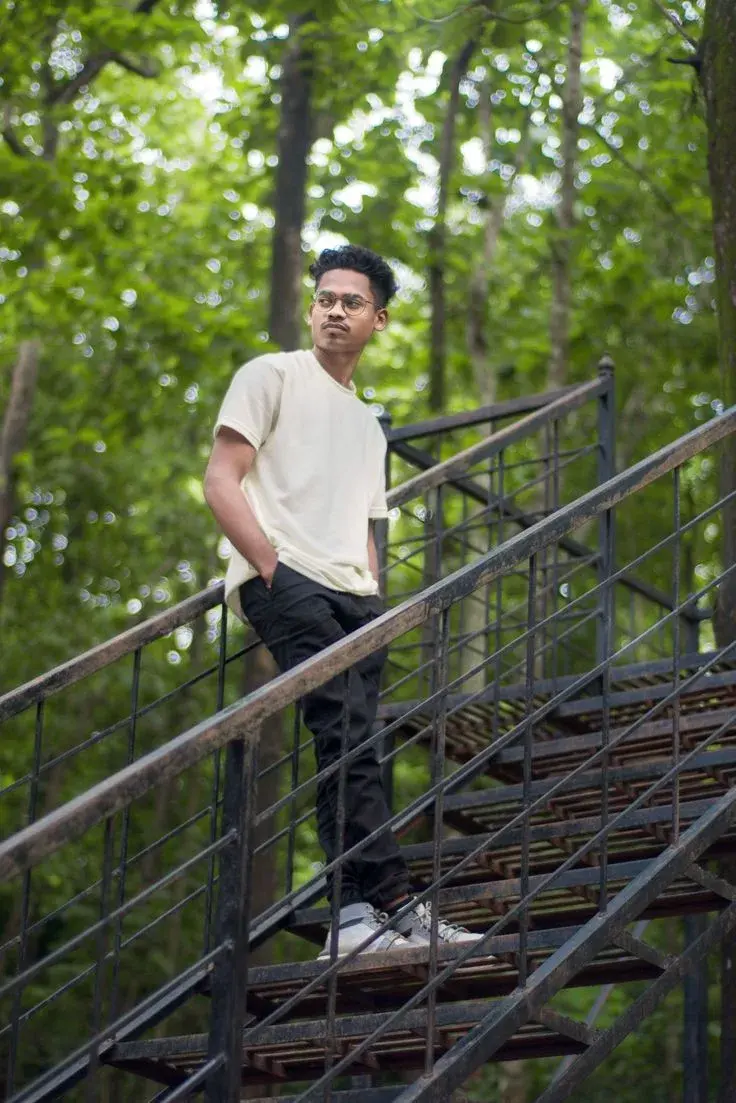 Candid Sunlit Forest Staircase Portrait Of Young South Asian Man With Glasses And T-Shirt Leaning On Metal Railing
