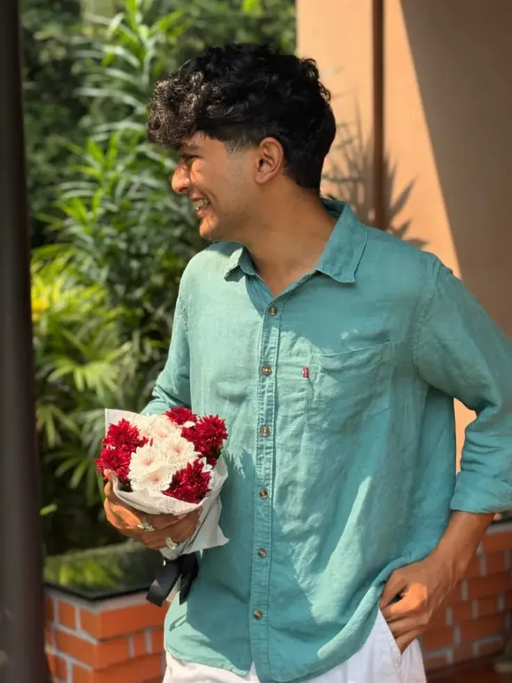 Candid Sunlit Garden Portrait Of Smiling Young South Asian Man With Curly Fringe Holding Bouquet Of Red And White Chrysa