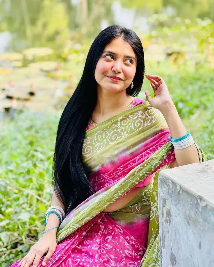 Candid Sunlit Garden Portrait Of Young South Asian Woman In Pink And Olive Batik Saree Leaning On Stone Ledge
