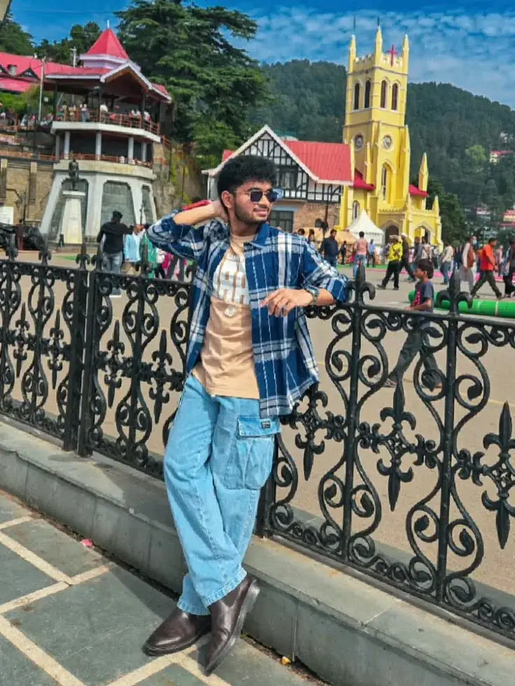 Candid Sunlit Heritage Church Promenade Portrait Of Young South Asian Man In Blue Plaid Shirt And Cargo Jeans Leaning On