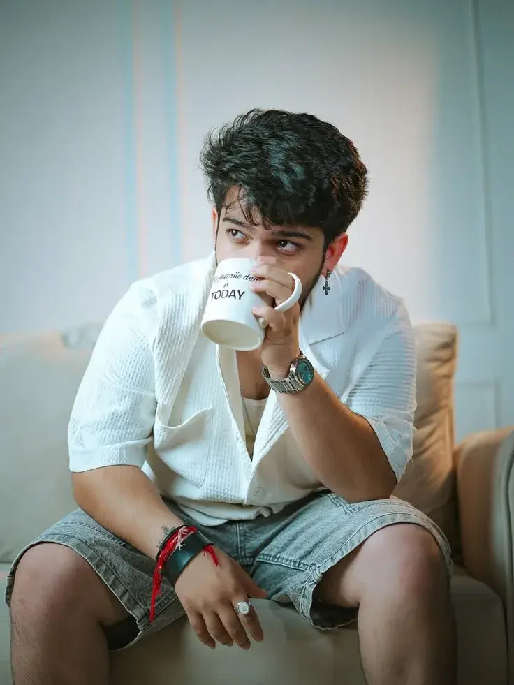Candid Sunlit Indoor Living Room Portrait Of Young South Asian Man Drinking Coffee On Beige Sofa In Textured White Shirt