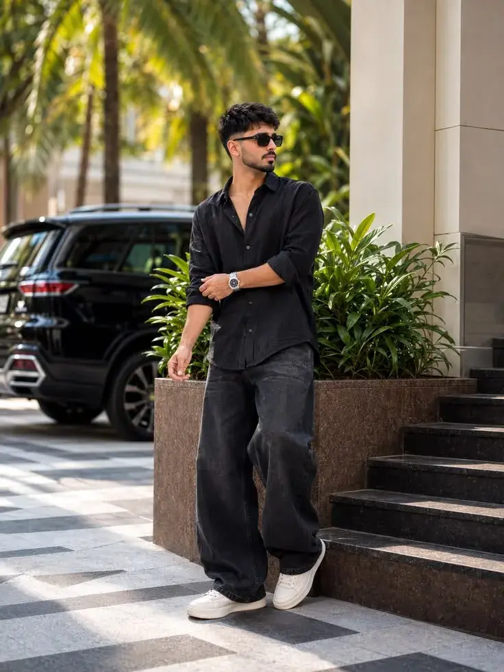 Candid Sunlit Luxury Villa Driveway Portrait Of Handsome Young South Asian Man In Black Linen Shirt Leaning On Stone Pla