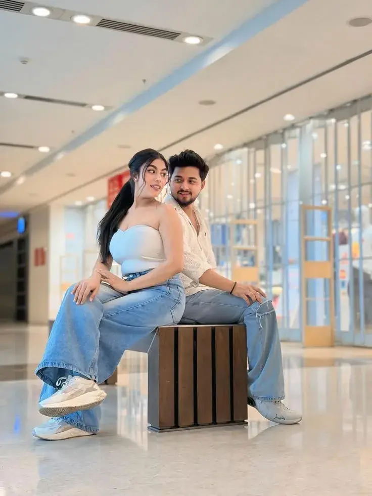 Candid Sunlit Mall Atrium Portrait Of Young South Asian Couple Seated Back-To-Back On Wooden Bench In Denim Jeans