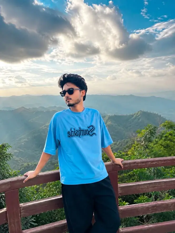 Candid Sunlit Mountain Viewpoint Portrait Of Young South Asian Man In Light Blue Graphic T-Shirt And Sunglasses Leaning
