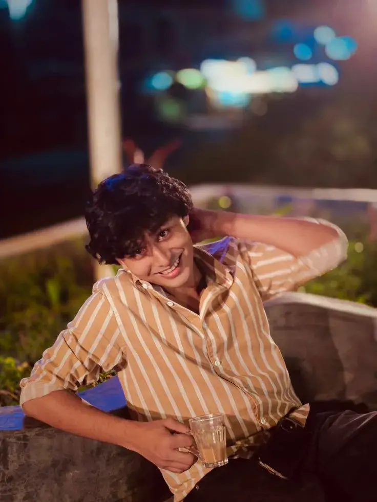 Candid Sunlit Outdoor Night Portrait Of Smiling Young South Asian Man With Curly Shag Hair In Striped Shirt Holding Glas