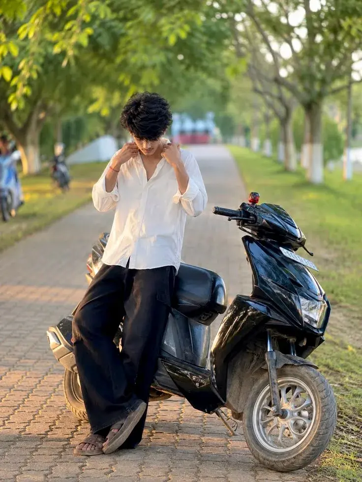 Candid Sunlit Park Promenade Portrait Of Young South Asian Man Adjusting White Linen Shirt Beside Black Scooter